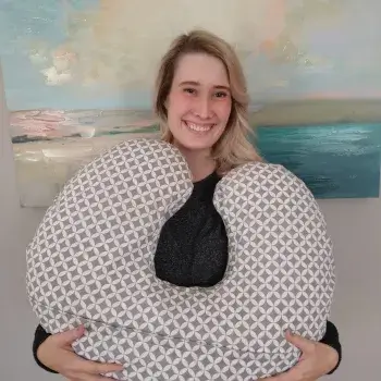 A person with blonde hair holding a gray and white patterned nursing pillow in front of a coastal-themed painting.