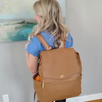 A person with long blond hair, wearing a blue shirt, is seen from the back carrying a large brown backpack. They are standing indoors near a wall with a framed painting.