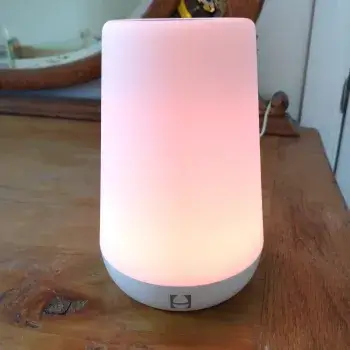 A small, cylindrical night light with a soft, warm pink glow is placed on a wooden surface.