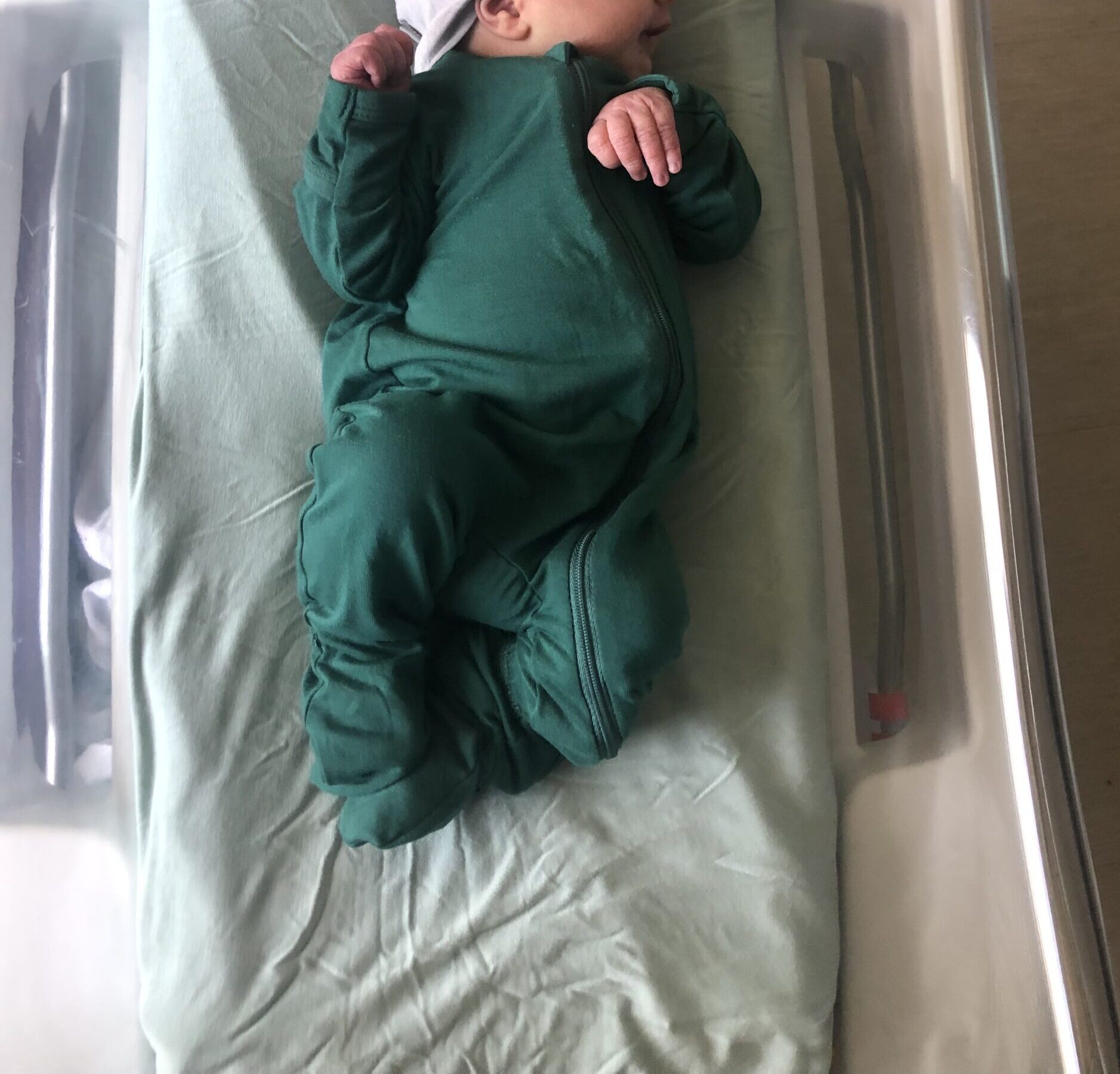 A newborn baby in a green outfit and gray hat sleeps in a hospital bassinet.