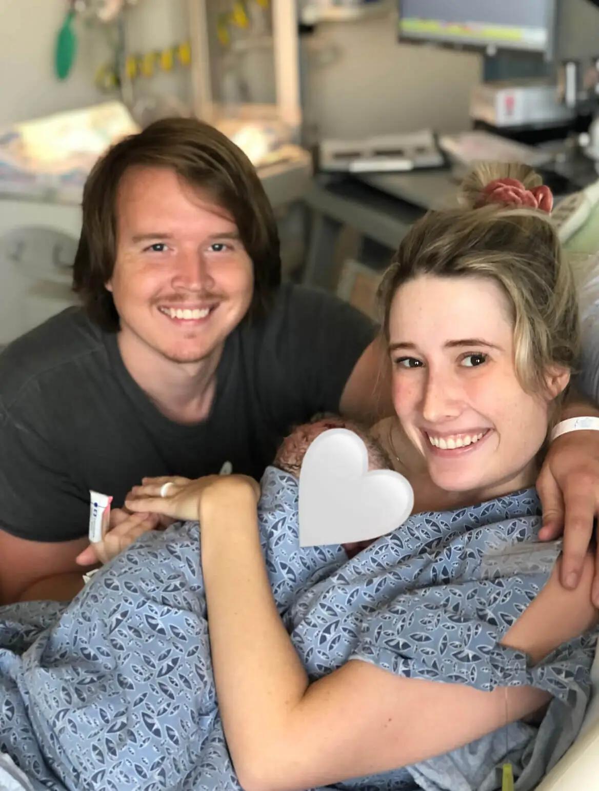 A smiling couple holds a newborn baby in a hospital room. The mother is in a hospital bed, holding the baby wrapped in a blanket.