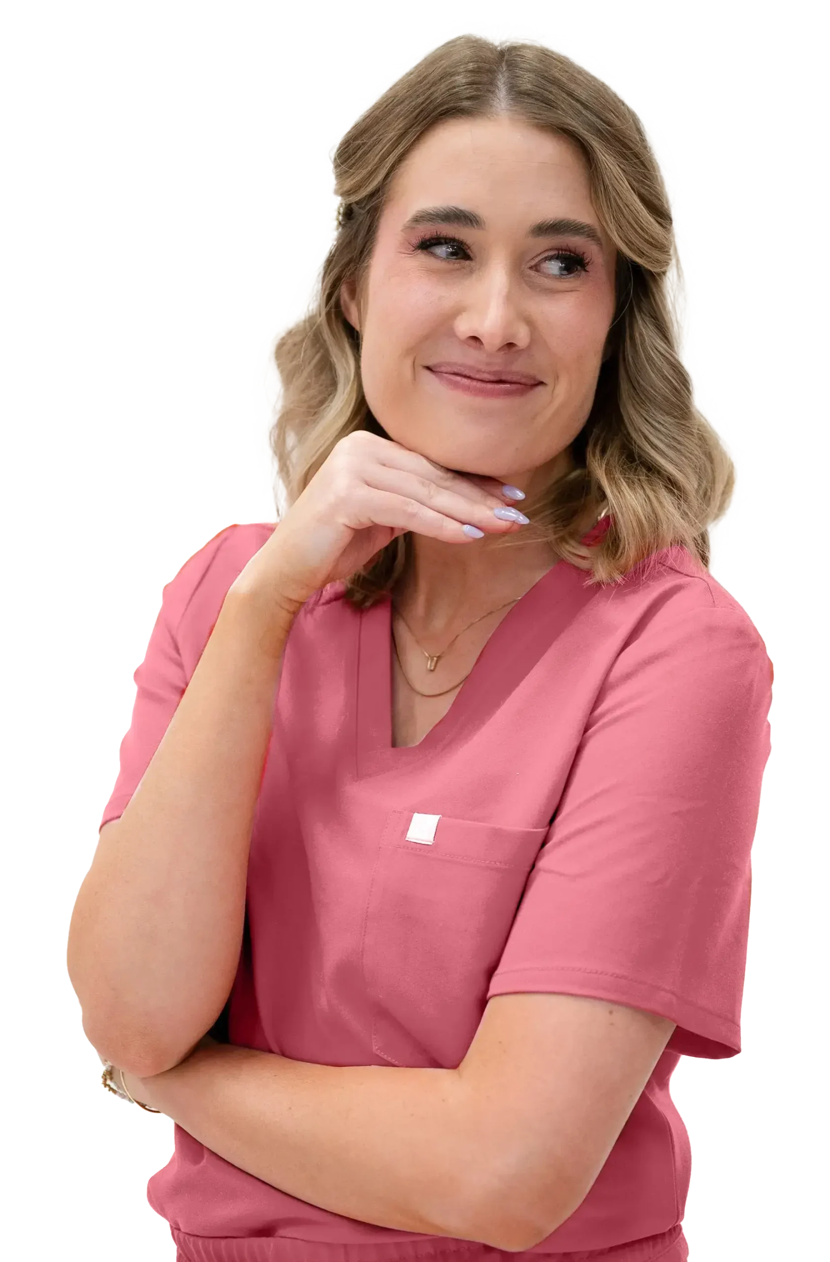 A woman in a pink scrub top stands with one arm crossed and her other hand under her chin, looking to the side and smiling.