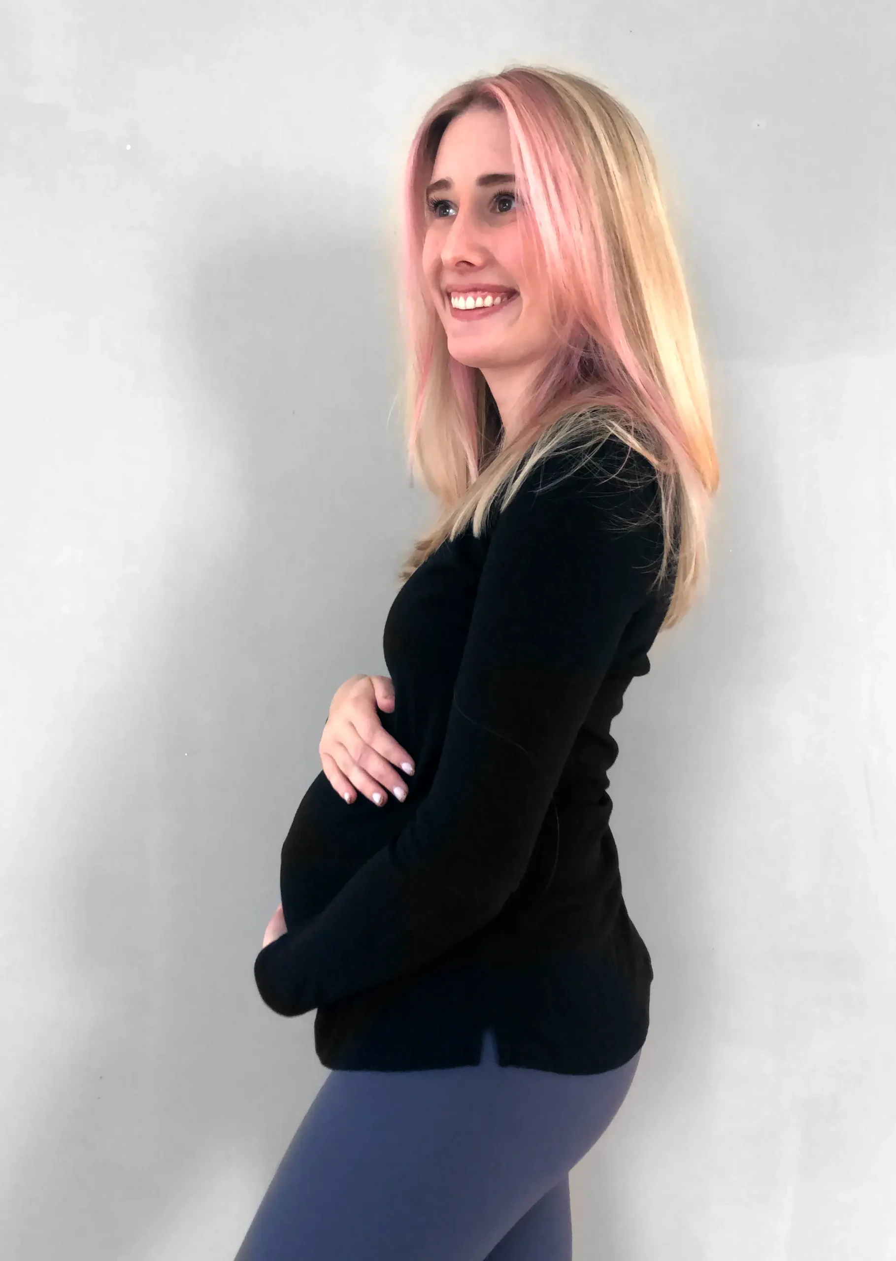 A woman with pink-tinted blonde hair smiles and holds her pregnant belly while wearing a black long-sleeve shirt and gray pants against a plain background.