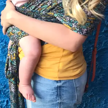 A woman wearing a yellow top and jeans carries a baby in a patterned sling against a blue textured background.