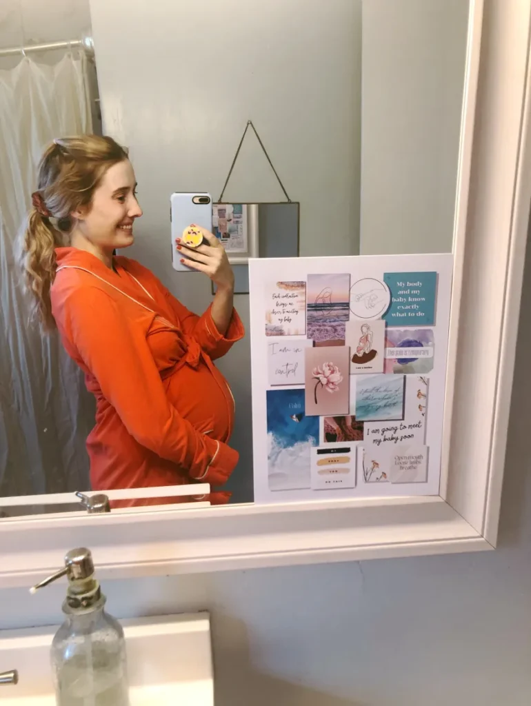 A woman in an orange robe takes a selfie in a bathroom mirror, showing her pregnant belly. The mirror is adorned with motivational quotes and images.