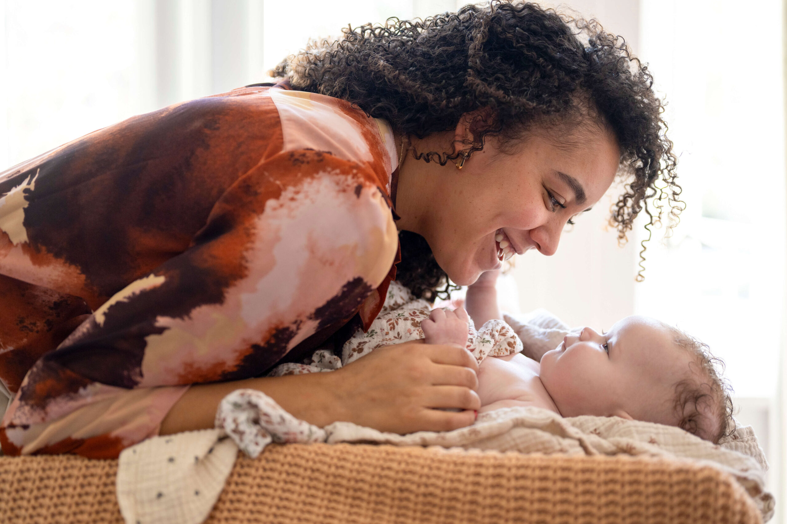 A woman with curly hair leans over and smiles at a baby lying in a woven basket. The baby, looking up, appears to be enjoying the interaction.