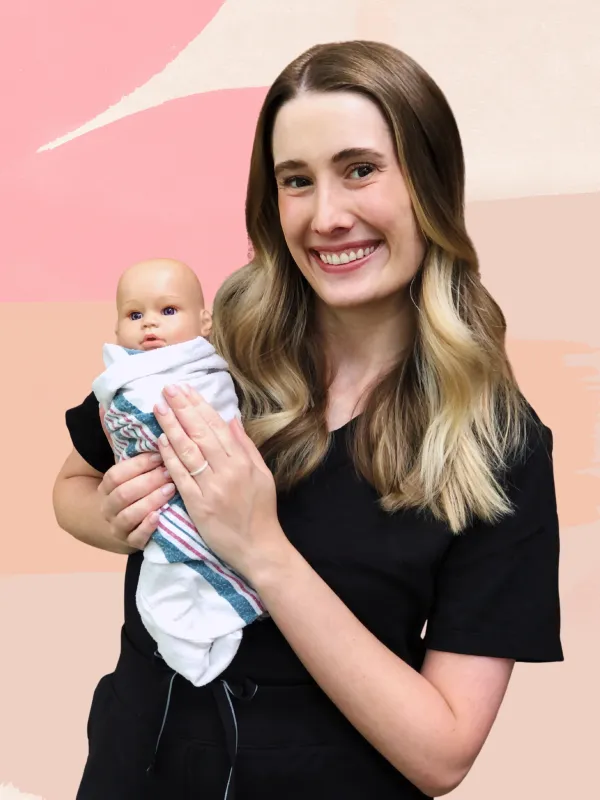 A woman with long hair smiles while holding a small baby doll wrapped in a blanket. She is standing against a pastel-colored background.