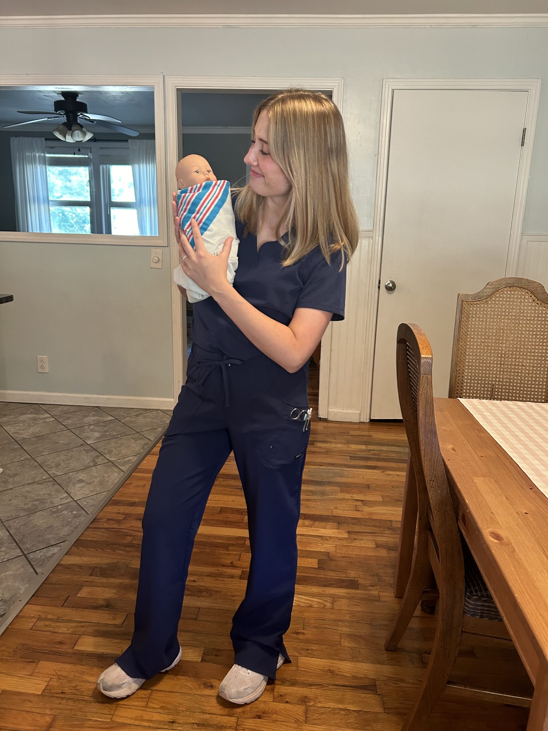 A person in blue scrubs holds a swaddled baby doll while standing in a kitchen area with wooden flooring and a dining table.